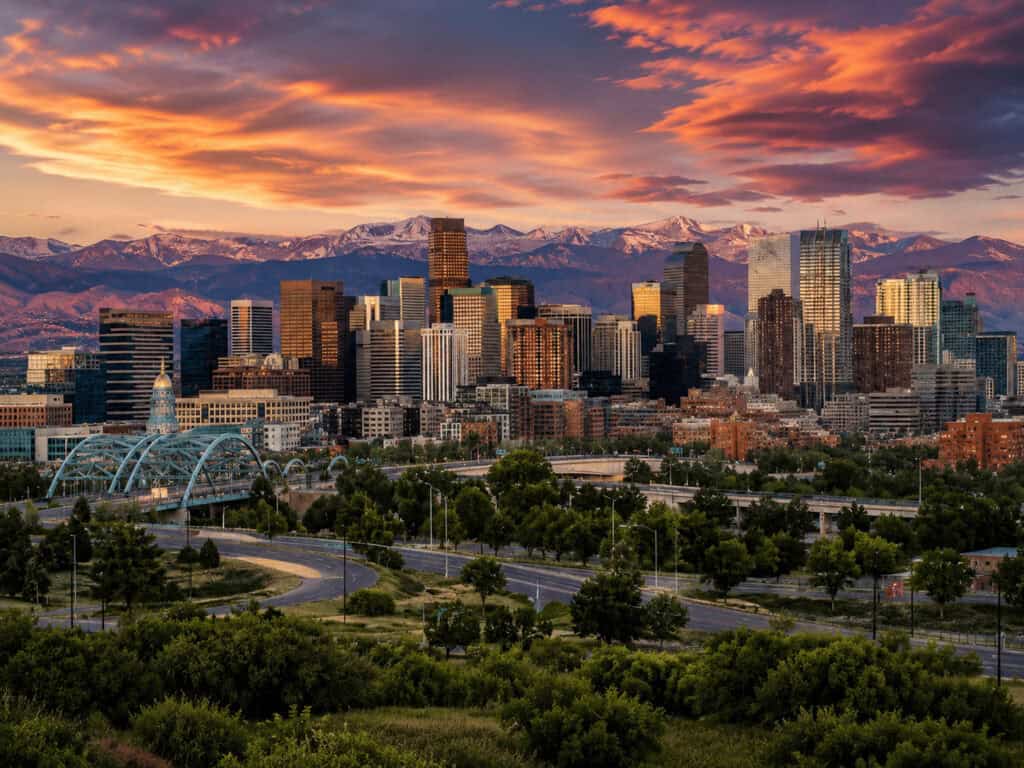 Denver skyline at sunset with mountains in the background and lush greenery in foreground.